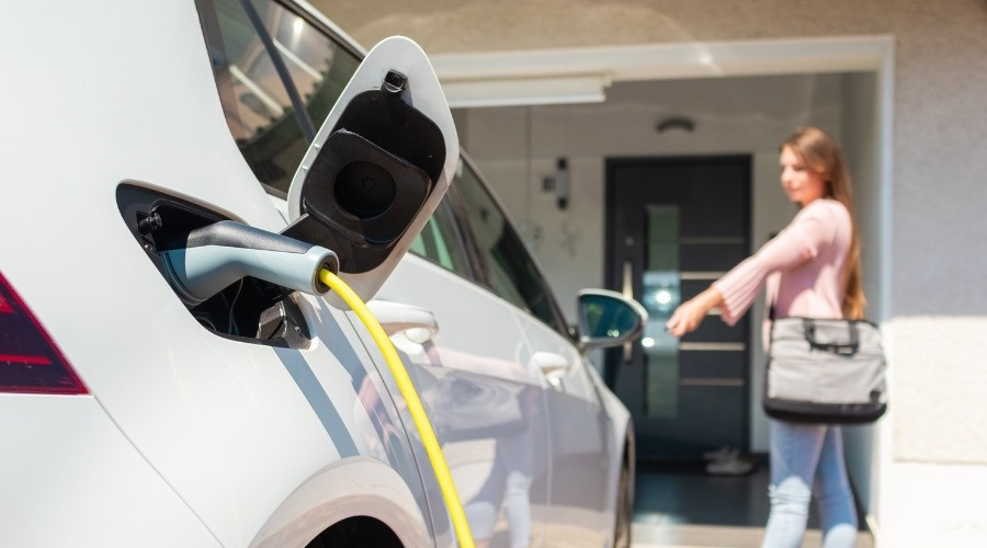 Electric vehicle charging station with a charging cable connected to a car, woman in the background preparing to leave, highlighting convenience and efficiency of home charging solutions.